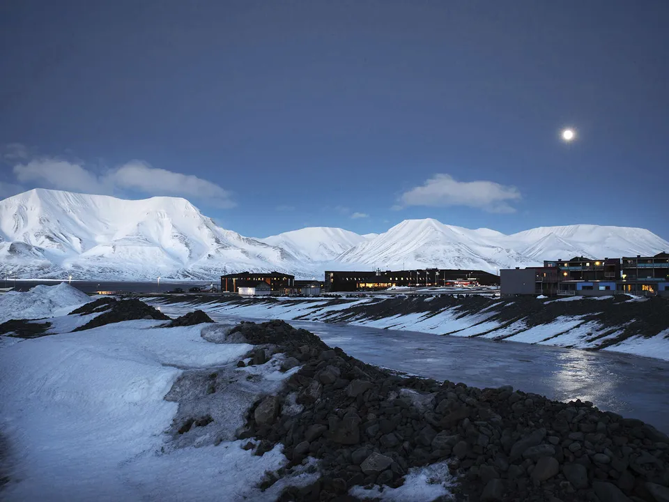 Alkutalven kuutamo valaisee maailman pohjoisimman asutuskeskuksen Longyearbyenin päivää. Keskellä kylää on
                              Norjan yliopistojen yhteinen tutkimuskeskus UNIS. Oikealla uusia hotelleja ja asuintaloja.